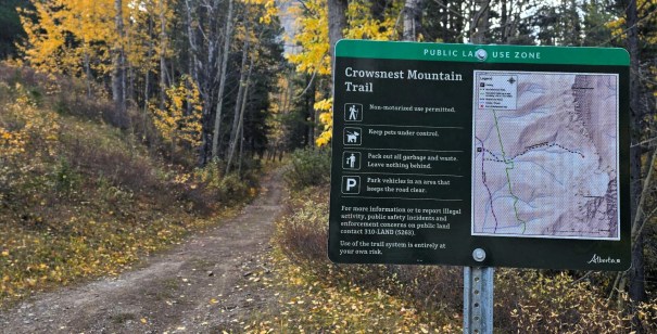 A sign for the Crowsnest Mountain Trail in a public land use zone, displaying rules and a map of the trail, surrounded by autumn foliage.