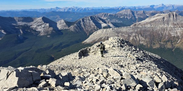 A person hiking on a rocky mountain summit with a panoramic view of a mountainous landscape and valleys in the background.