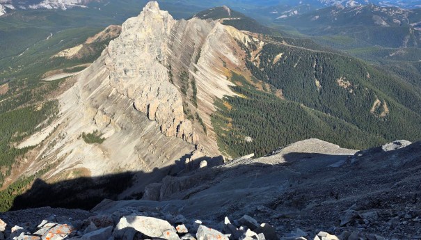 A panoramic view of a rugged mountain peak with distinct geological layers, surrounded by lush green forests and distant hills under a clear blue sky.