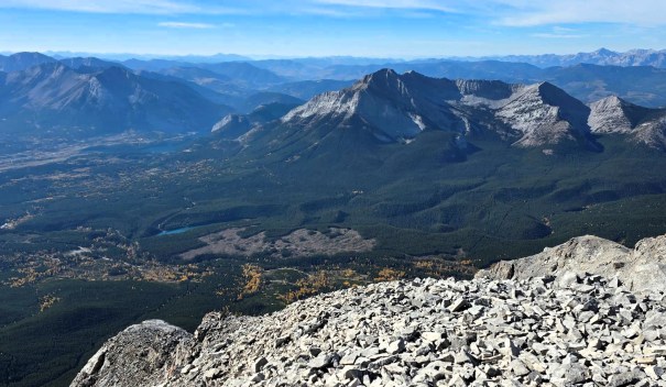 A panoramic view of mountainous terrain with rocky peaks, lush green valleys, and a clear blue sky in the background.