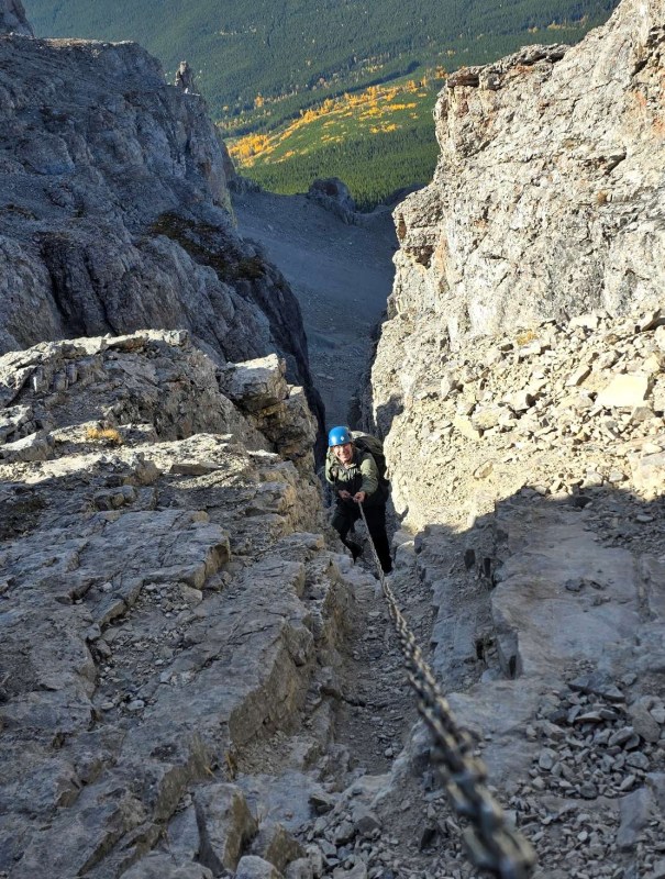 A climber ascending a rocky cliff using a chain for support, surrounded by mountainous terrain and autumn foliage in the background.