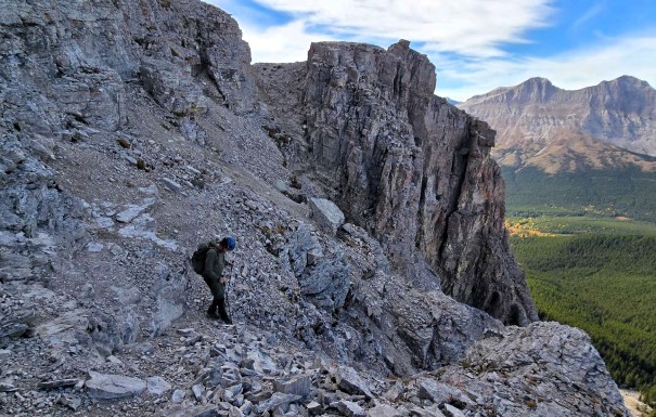 A hiker navigating a rocky mountain path with steep cliffs and a forested valley below, under a partly cloudy sky.