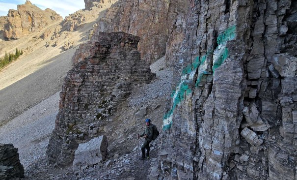 A hiker standing on a rocky trail beside a steep cliff with green markings on the rock, surrounded by mountainous terrain.