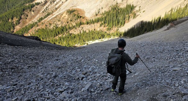 A hiker navigating a rocky slope in a mountainous area, surrounded by coniferous trees and steep terrain.