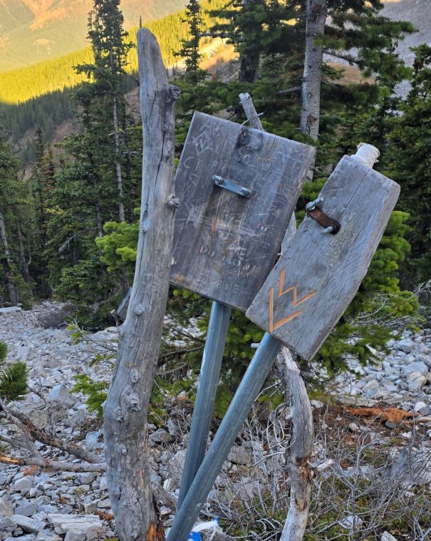 Two weathered wooden signs mounted on metal posts, surrounded by trees and rocky terrain.